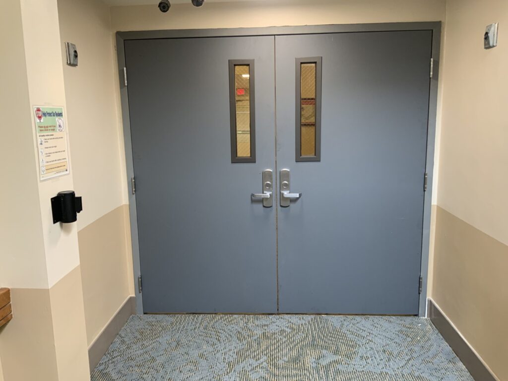 Closed double blue doors with rectangular windows in a hallway, beige walls, and blue-patterned carpeted floor.