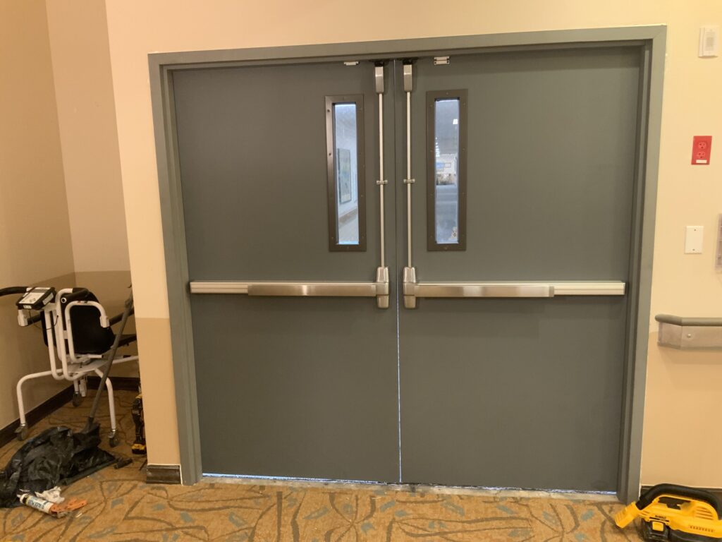 Gray double doors with push bars and small windows, set in a beige wall, with tools and a chair nearby.