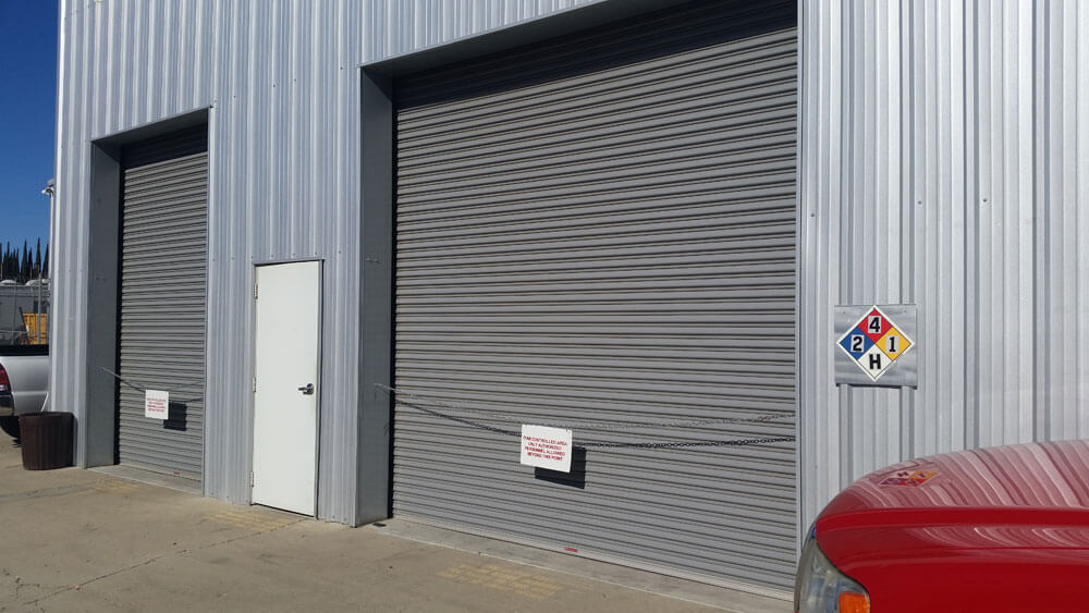 Two closed metal roll-up doors and a white door on a corrugated building, with a hazard sign and warning signs visible.