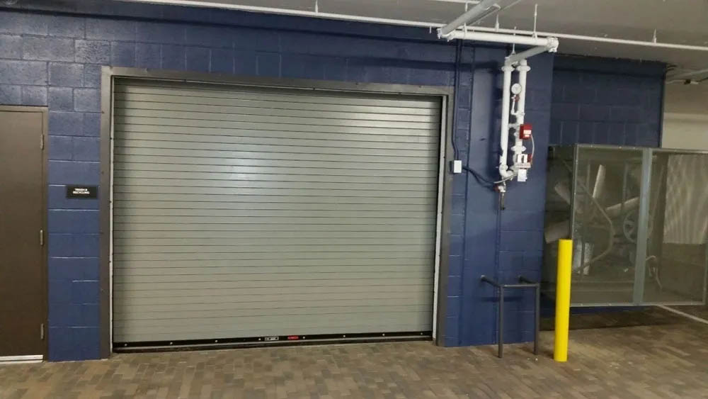 A closed gray roll-up garage door set in a blue wall, with a nearby yellow bollard and utility pipes above.