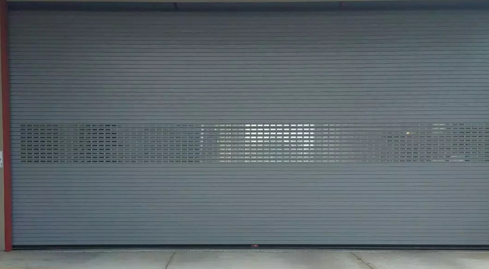 Closed gray metal roller shutter door with a horizontal row of perforated vents in the middle. Concrete ground visible.